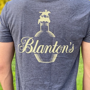 Puede incluir: Una camiseta gris oscura con un gráfico amarillo de una botella con un caballo en la parte superior y el texto "Blanton's" en una fuente cursiva.