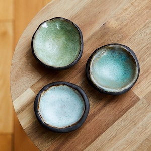 May include: Three small, handmade ceramic bowls with a dark rim and a textured, glazed interior. The bowls are in shades of green, blue, and white. They are arranged on a round wooden surface, showcasing their unique colors and textures.