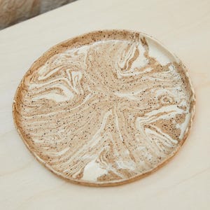 May include: A round ceramic plate with a marbled design in shades of beige and white. The plate has a slightly raised edge and is speckled with small black dots. The plate is set on a light-colored wooden surface.