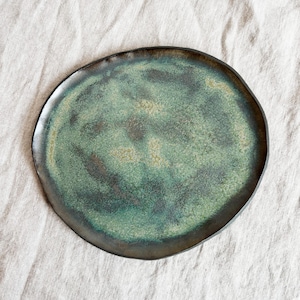 May include: A handmade ceramic plate with a unique, organic shape. The plate features a mottled green and brown glaze, with a dark brown rim. The surface has a speckled texture, adding to its artisanal quality. The plate is a stylish addition to any kitchen.