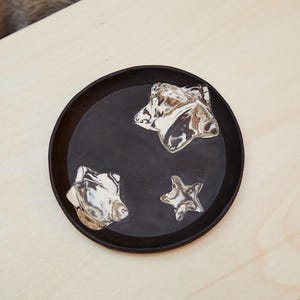 May include: A round, black ceramic tray with three silver star-shaped embellishments. The tray is set against a light wood surface, creating a contrast between the dark and light tones.