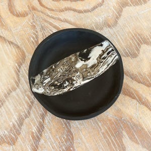 May include: A small, round, black ceramic dish with a decorative, oblong, textured accent piece in shades of white, brown, and black. The dish is set on a light-colored, wood-grain surface.