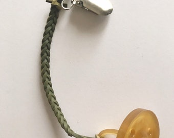 Può includere: Un ciuccio per bambini con tettarella e scudo in gomma naturale, collegato a un cinturino intrecciato verde oliva e a una clip in metallo. Il ciuccio è di colore ambra chiaro.