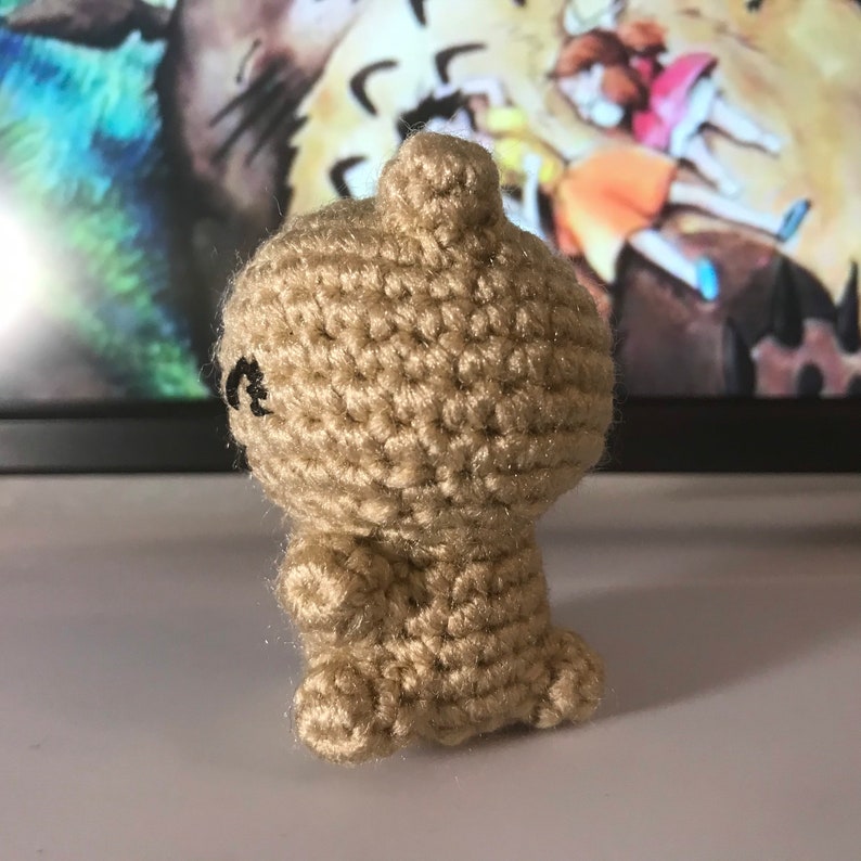 May include: A small, tan crocheted figure with a rounded body, small arms, and feet. The figure has a small, rounded head and black embroidered eyes. The yarn has a slight shimmer. The figure is likely a handmade toy or decorative item.