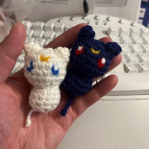 May include: Two crocheted cat figures, one white and one navy blue, held in a hand. The white cat has yellow and blue accents, while the blue cat has red and yellow accents. Both have small tails.