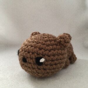 May include: A small, handmade brown crochet bear with black eyes and a white highlight. The bear is lying down, with its small ears visible. The toy is made of yarn and has a textured appearance.