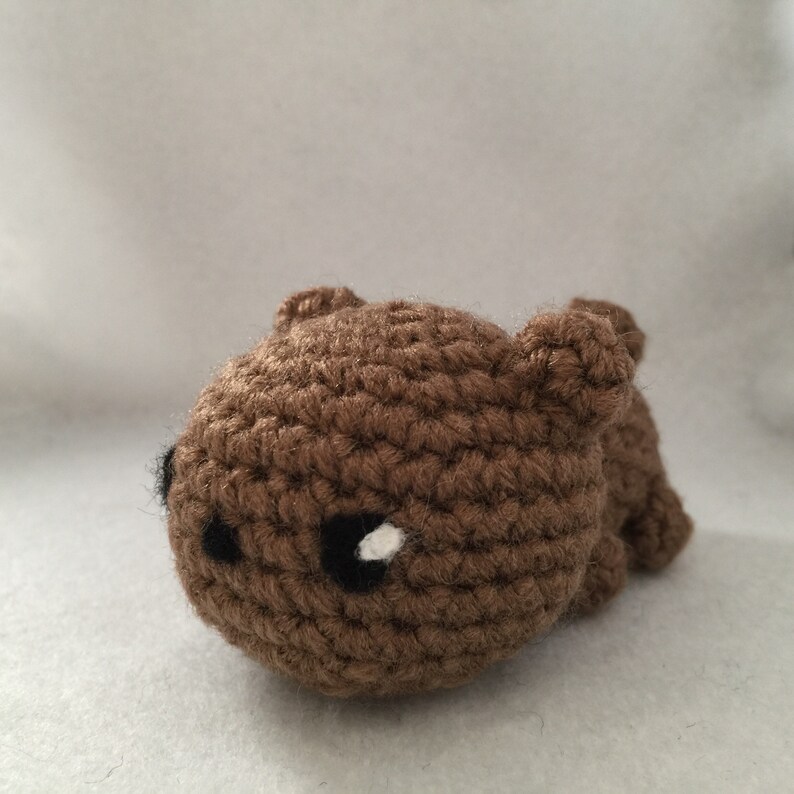 May include: A small, handmade brown crochet bear with black eyes and a white highlight. The bear is lying down, with its small ears visible. The toy is made of yarn and has a textured appearance.