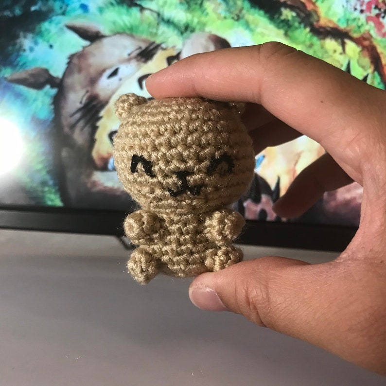 May include: A small, tan crocheted bear with black embroidered facial features. The handmade bear has a rounded body, small arms and legs, and a smiling face. The bear is held in a person's hand.