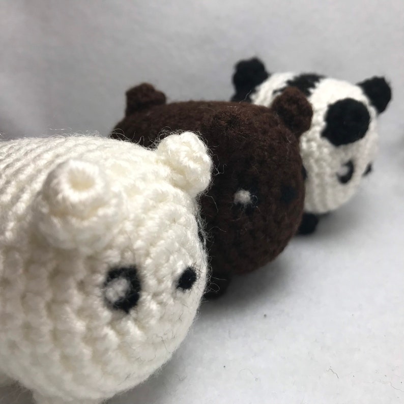 May include: Three crocheted bear figurines in a row. The first is white, the second is brown, and the third is a panda bear with black and white markings. Each bear has small black eyes and a cute, rounded shape.