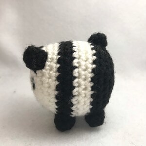 May include: A handmade, crocheted panda bear with black and white stripes. The small stuffed animal has black ears, eyes, and feet. The panda is a round shape and is sitting on a white surface.