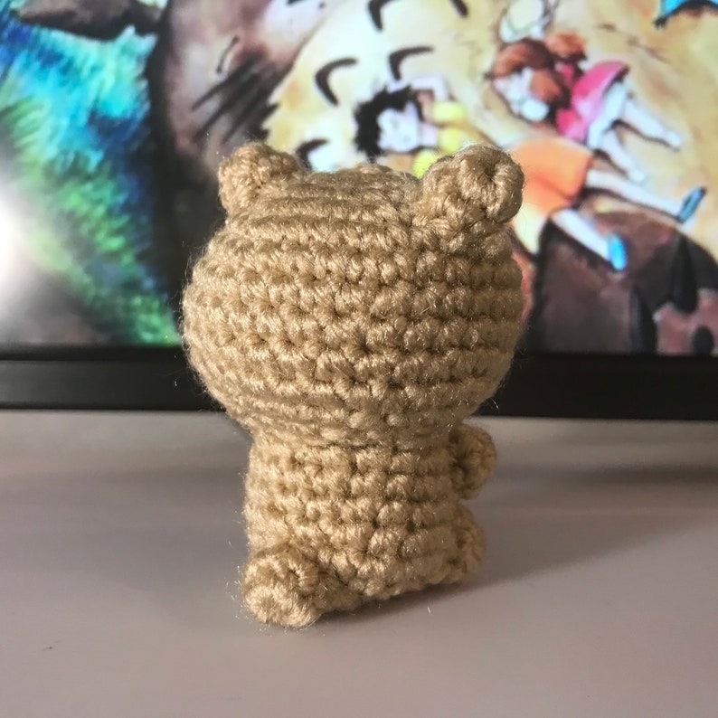 May include: A small, tan crocheted bear figurine. The bear has a rounded body, small ears, and tiny arms and legs. The yarn is a solid, light brown color. The bear is facing away from the camera.