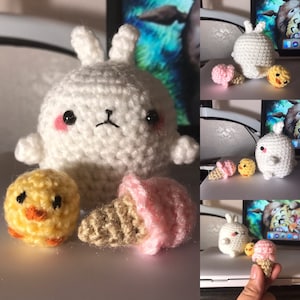 May include: A collection of handmade crochet toys, including a white bunny with pink cheeks, a yellow chick, and a pink ice cream cone. The toys are displayed on a white surface, with a computer screen in the background.