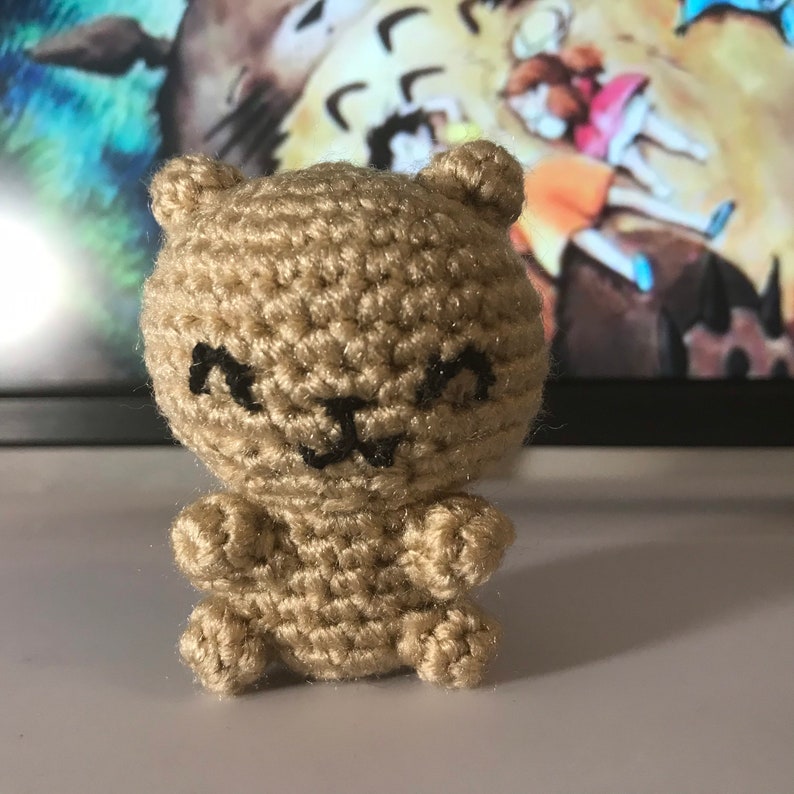 May include: A small, handmade crochet bear figurine in a light tan color. The bear has a smiling face with black embroidered details. The bear is sitting upright with small arms and legs. The background is blurred.
