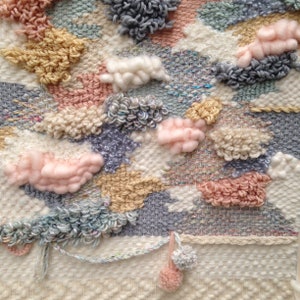 May include: A woven wall hanging with a textured surface and various colours, including pink, blue, and beige. The hanging features a variety of shapes and textures, including clouds, swirls, and pom-poms.