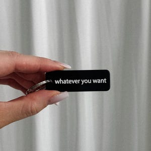 May include: Black rectangular keychain with the phrase "whatever you want" in white lettering. The keychain is held in a hand against a white curtain backdrop.