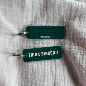 May include: Two rectangular, dark green keychains with silver key rings. One keychain has the text "4headkiss" in white. The other keychain has the text "THINK BIGGER!!" in white. The keychains are on a white, textured surface.