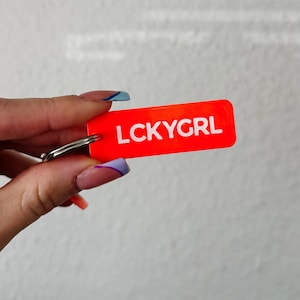 May include: A bright orange rectangular keychain with the text "LCKYGRL" in white. The keychain is attached to a silver metal ring.