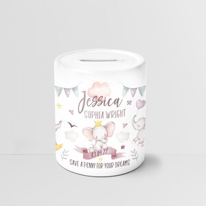 Può includere: Salvadanaio in ceramica bianca con un design di elefante rosa. Il salvadanaio ha una nuvola rosa con il nome "Jessica Sophia Wright" e la data "03.09.22". Il testo "Save a penny for your dreams" è stampato sul fondo.
