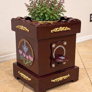 May include: A dark brown wooden planter with gold accents and a decorative floral design. The planter has a scalloped top and a small shelf. A green plant with purple flowers sits inside the planter.