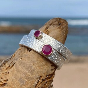 Puede incluir: Dos anillos de plata con bandas texturizadas y gemas redondas de color rojo intenso. Los anillos están apilados, con una gema más grande que la otra. Los anillos se exhiben sobre un trozo de madera flotante, con el océano al fondo.