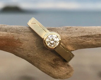 May include: A simple gold band ring with a single round diamond set in a bezel setting. The ring is resting on a piece of driftwood.
