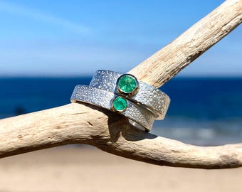 May include: Two silver rings with textured surfaces and emerald gemstones. The rings are stacked on a piece of driftwood, with a blue ocean and sky in the background. The emeralds are round and set in gold bezels.