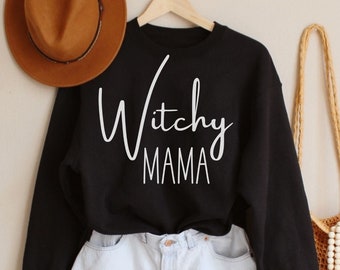 May include: A black sweatshirt with the text "Witchy Mama" in white script is displayed with light blue denim shorts, a brown hat, and a woven tote bag. The items are arranged against a neutral background, creating a casual, stylish aesthetic.