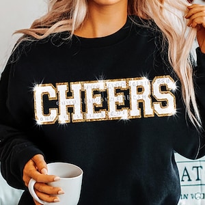 May include: Black sweatshirt with the word "CHEERS" in large, white, sparkly letters. The letters are outlined in gold. The person in the image is holding a white mug and wearing blue jeans.