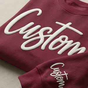 May include: A maroon sweatshirt and sleeve with the word "Custom" in white script. The sweatshirt is folded, and the sleeve is visible below. The design is a custom apparel item.