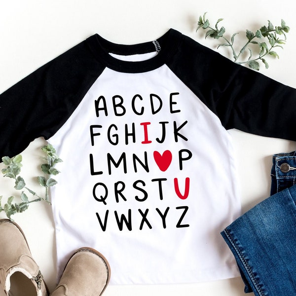 以下が含まれることがあります： アルファベットがプリントされた白と黒のラグランスタイルのTシャツ。 「I」と「U」の文字は赤色で、間に赤いハートがあります。 シャツは青いジーンズとベージュのブーツと組み合わせています。