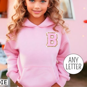 May include: Pink hoodie with a gold glitter letter B on the front. The hoodie has a large pocket on the front. The text "ANY LETTER" is visible in the bottom right corner of the image.