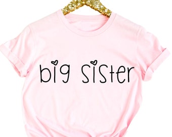 Può includere: Maglietta rosa con il testo "big sister" in lettere nere e due piccoli cuori sopra il testo.