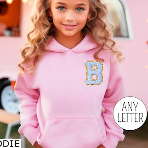 May include: Pink hoodie with a light blue letter B with gold glitter on the front. The text "ANY LETTER" is visible in the bottom right corner of the image. The word "HOODIE" is visible in the bottom left corner of the image.