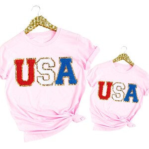 May include: Two pink t-shirts with the word "USA" in red, white, and blue glitter letters. The shirts are on hangers.