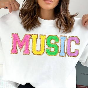 Puede incluir: Sudadera blanca con la palabra "MUSIC" en letras de colores y peludas. Las letras están delineadas en oro.