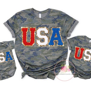 May include: Matching camouflage t-shirts with the word "USA" in red, white, and blue glitter letters. The shirts are in different sizes, one for an adult and one for a child.