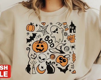 以下が含まれることがあります： ハロウィーンをテーマにしたデザインのベージュのスウェットシャツ。カボチャ、幽霊、コウモリ、そして「BOO」の文字が含まれています。スウェットシャツはクルーネックと長袖です。「FLASH SALE」という赤いバナーが見えます。