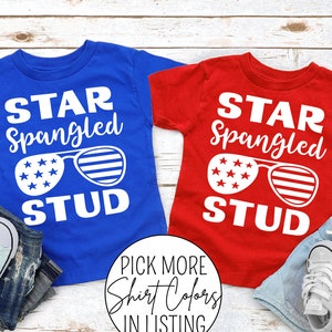 May include: Two red and blue t-shirts with the text "Star Spangled Stud" and a graphic of sunglasses with stars and stripes. The shirts are laid out on a white wood background with American flags and sneakers.