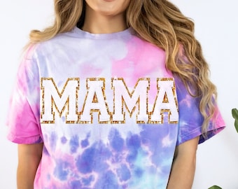 Op de afbeelding: Een tie-dye T-shirt met een roze, blauwe en witte kleurstelling. Het shirt heeft het woord "MAMA" in witte letters met een gouden glitterrand.