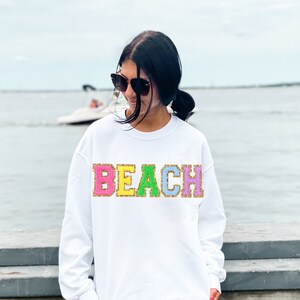 May include: White sweatshirt with the word "BEACH" in colorful glitter letters. The sweatshirt is worn by a person standing on a wooden dock.