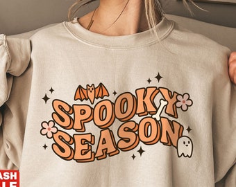 Könnte beinhalten: Beiges Sweatshirt mit den Worten "Spooky Season" in Orange und Weiß, mit einer Fledermaus, einem Knochen, Blumen und einer Geistergrafik. Das Sweatshirt hat lange Ärmel und einen Rundhalsausschnitt. Ein rotes "Flash Sale" Banner ist sichtbar.