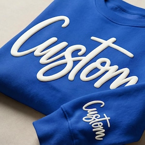 May include: A folded royal blue sweatshirt with the word "Custom" in white, cursive lettering across the front. The sleeve cuff also displays the word "Custom". The sweatshirt is made of a soft material.