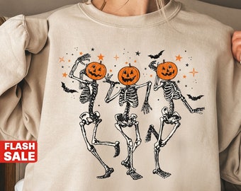May include: A sand-coloured sweatshirt with a Halloween-themed design. The graphic shows three dancing skeletons with pumpkin heads, bats, and stars. The text "SAND SWEATSHIRT" is at the bottom of the image.