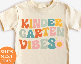Puede incluir: Camiseta color crema con las palabras "KINDERGARTEN VIBES" en letras coloridas de estilo retro. Un pequeño gráfico de flores está al lado de la palabra "VIBES". La camiseta es de cuello redondo y manga corta.