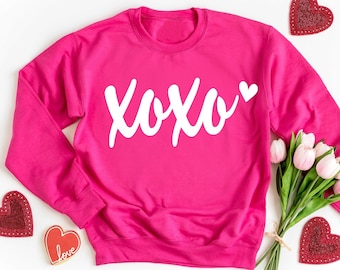 May include: A bright pink sweatshirt with the word "XOXO" in white script. The sweatshirt is surrounded by Valentine's Day decorations, including heart-shaped decorations and a bouquet of tulips.