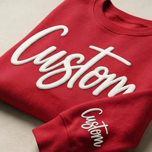 May include: A folded red sweatshirt with the word "Custom" in white, cursive lettering across the front. The sleeve of the sweatshirt is also visible, with the same word in a smaller size.