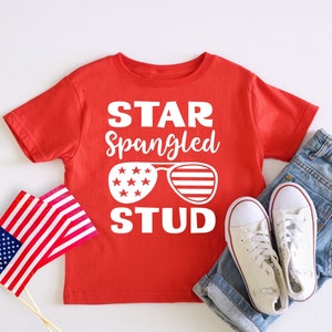 May include: Red short-sleeved t-shirt with a white graphic that says "Star Spangled Stud" with a pair of sunglasses with stars and stripes. The sunglasses are white with red and blue accents.