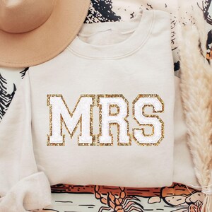 May include: A cream-colored sweatshirt with the word "MRS" in white letters outlined in gold glitter.