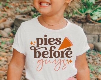May include: A young child wearing a white t-shirt with the words "pies before guys" in brown and pink script, and a slice of pie graphic. The child is smiling and wearing a pink headband and pink shorts.
