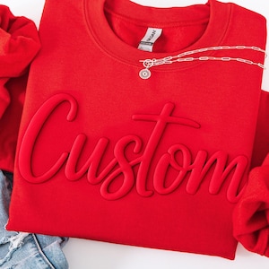 May include: A bright red long-sleeve sweatshirt featuring the word Custom embossed in a matching red raised script font across the chest. A silver-toned chain necklace with a small circular pendant rests on the neckline of the garment.